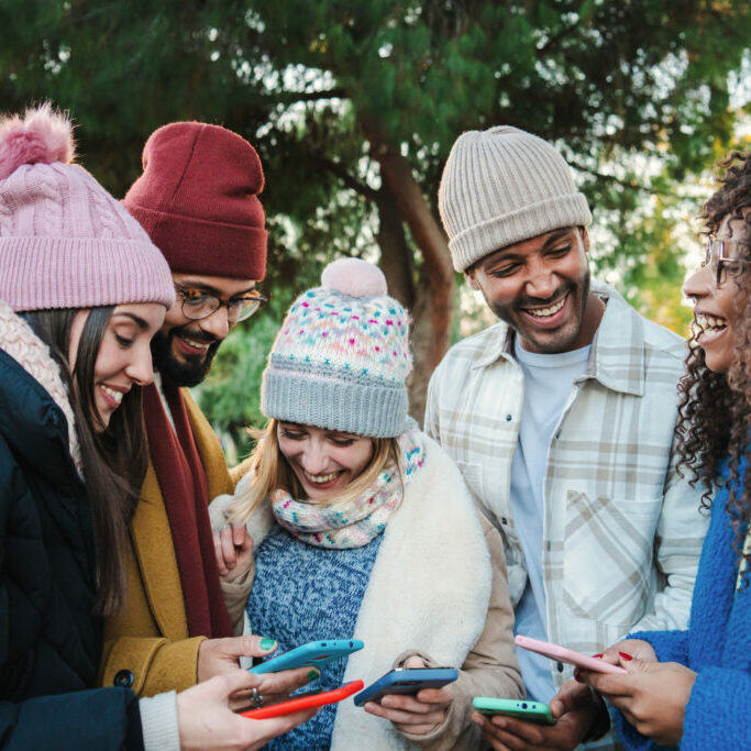 Group of multiracial young friends with coats and hats, smiling and watching the social media with a cellphone app. Portrait of happy people having fun sharing media and laughing with a smart phone. Communication concept. High quality photo